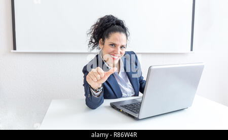 Ritratto di un normale business donna facendo pollice in alto segno in una scrivania in ufficio. Con copia spazio. Vestirsi in modo professionale sorridente e guardando la telecamera. Capelli neri e occhi blu Foto Stock