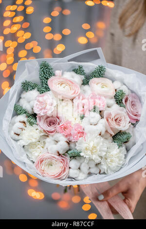 Inverno luminoso bouquet di fiori misti con cotone, rami di pino Nobilis e Ranunculus. Mazzetto floreale con ranuncolo persiano nelle mani della donna. Garland bokeh sullo sfondo. Amore di natale Foto Stock