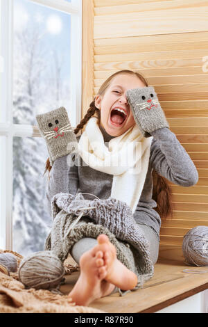 Bambino ragazza è seduta su un davanzale e giocare con il guanto fatti a mano. Bella vista al di fuori della finestra - giornata di sole in inverno e neve. Foto Stock