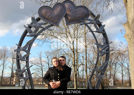 Amare giovane nel giorno di San Valentino in un parco della città contro un lago sotto un grande cuore forgiato Foto Stock