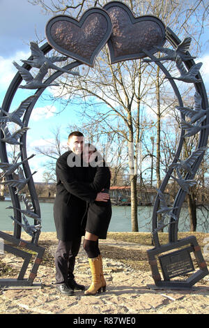 Amare giovane nel giorno di San Valentino in un parco della città contro un lago sotto un grande cuore forgiato Foto Stock