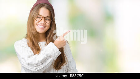 Giovane bella bruna hipster donna che indossa occhiali e cappello invernale su sfondo isolato allegro con un sorriso della faccia rivolta con la mano e fi Foto Stock