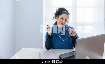 Ritratto di una donna normale con i capelli neri e la pelle bianca guardando la telecamera e tenendo gli occhiali dalla sua scrivania. In un ufficio con un ordine del giorno, laptop e notebook Foto Stock