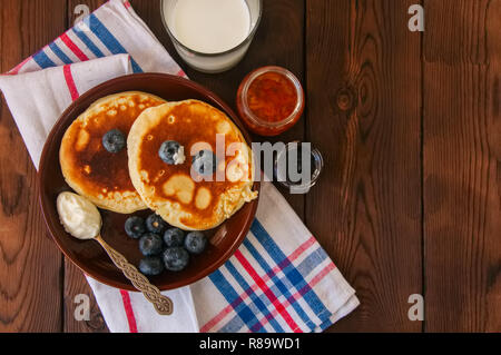 Vista superiore del Pancake fatti in casa in una piastra, latte in un bicchiere, marmellata su una cornice di legno. Stile rustico. Foto Stock