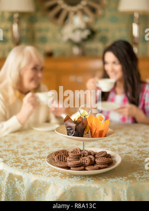 Marrone deliziosi biscotti e tortine In luminose sono di incarto sul cavalletto di dessert al lato di una tabella. Immagine sfocata del Senior madre parlando con H Foto Stock