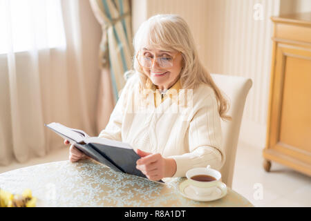 Affascinante la nonna legge un libro interessante di bere un tè Warn. Concetto di pensionamento Foto Stock