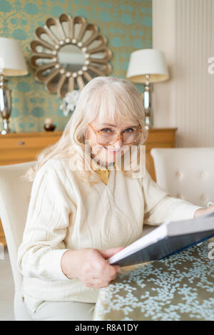 Ritratto su una bella vecchia signora con gli occhiali per leggere un libro. Elegante casa e la nonna in bicchieri. Foto Stock