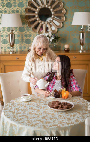 Trascorrere del tempo insieme. Sorridente Madre Senior versando il tè di porcellana teiera in la figlia della tazza. Biscotti e tortine sono sul Tabl Foto Stock