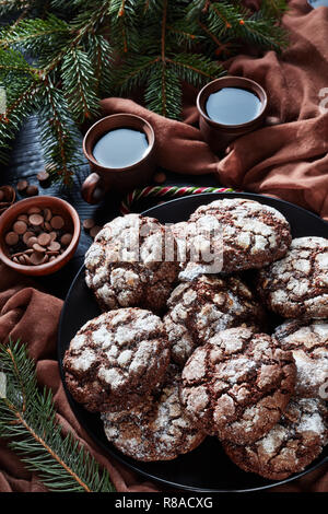 Close-up di Natale Chocolate Crinkle cookies su una piastra su un nero tavolo in legno di cipresso, candy canes, panno marrone e tazze di caffè, rustico Foto Stock