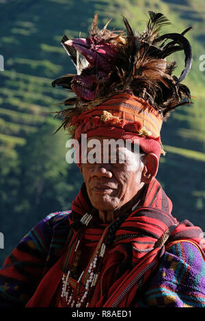 Un anziano uomo Ifugao indossare copricapo ornato con piume e monkey cranio al Banaue terrazze di riso nel nord di Luzon, Filippine. In passato gli Ifugao noto anche come Ifugaw, Ipugao, Yfugao temevano erano cacciatori di teste, così come altre tribù delle regioni montuose del Luzon del nord. Foto Stock