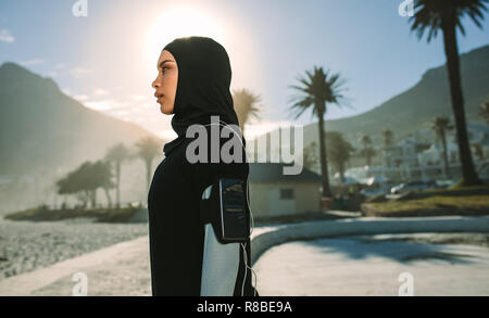 Vista laterale del hijab giovane donna in piedi all'aperto con la luce solare diretta dal retro. Donna musulmana riposo dopo una corsa mattutina nella città. Foto Stock