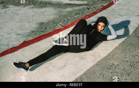 Sport donna in hijab facendo bicicletta crunch allenamento all'aperto. Femmina sdraiato sul pavimento di cemento e abs facendo esercizi di allenamento in mattinata. Foto Stock