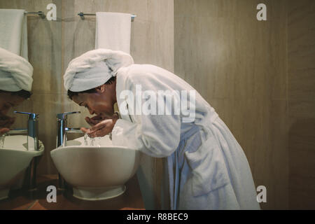 Donna africana in accappatoio lavando la faccia nel lavandino del bagno. Femmina matura il lavaggio il suo volto in bagno. Foto Stock