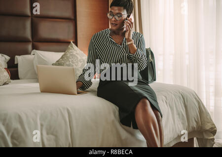 Donna africana in hotel moderno camera seduta sul letto tramite laptop e parlando su telefono cellulare. Business Woman in camera in hotel utilizzando computer portatile e lo smartphone Foto Stock