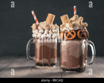 Vista ravvicinata dei due freakshake a mason jar sul tavolo marrone. Freaked frullato al cioccolato, pretzel, marshmallow, popcorn e waffles. Cibo alla moda Foto Stock