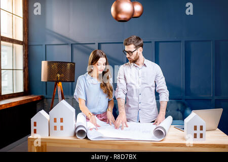 Giovane donna e uomo come gli architetti che lavorano con blueprint, laptop e modelli della casa in ufficio Foto Stock