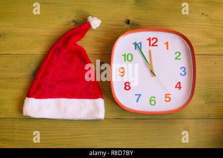 Immagine di Santa Hat e un orologio che mostra cinque minuti a mezzanotte su un tavolo di legno Foto Stock