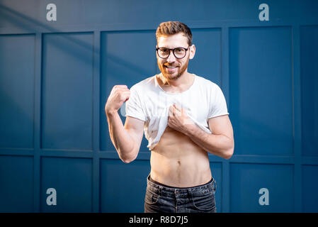 Ritratto di un bel uomo forte che mostra e bicipiti muscoli addominali sulla parete blu sullo sfondo Foto Stock