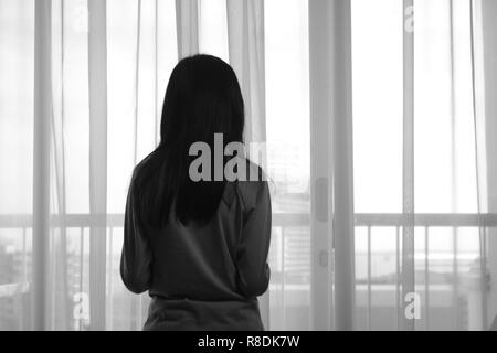 Vista posteriore di immagine triste ragazza solitaria guardando attraverso la finestra in bianco e nero la foto in bianco e nero Foto Stock