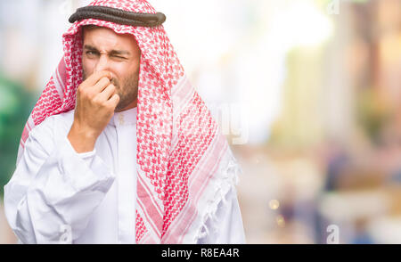 Giovane uomo bello indossare keffiyeh su sfondo isolato annusare qualcosa di maleodorante e disgustoso, intollerabile odore, trattenendo il respiro con le dita Foto Stock