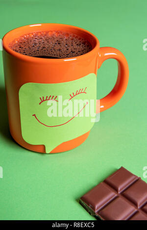 Il caffè caldo in una tazza di colore arancione e una divertente Smiley face nota adesiva su un tavolo verde accanto a un grande pezzo di cioccolato Foto Stock