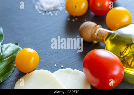 Il giallo e il rosso dei pomodori, mozzarella, olio di oliva e il verde del basilico sono giacenti in cerchio su scuro scheda di scisto. Foto Stock