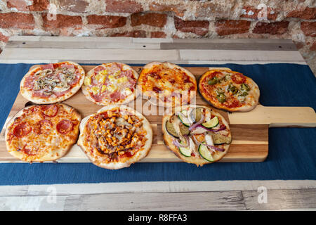 Sette piccole pizze su una tavola di legno, freschi di forno, pronto a mangiare in un ristorante. Su un tavolo di legno con una tovaglia blu e un muro di mattoni Foto Stock