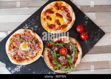 Vista aerea di tre diverse pizze su una lavagna nera guarnire con farina e due cherry pomodoro su una tavola di legno tavolo bianco Foto Stock