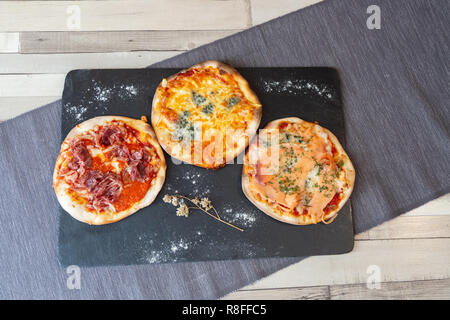 Vista aerea di tre diverse pizze su una lavagna nera guarnire con farina e fiore secco su una tavola di legno tavolo bianco grigio di una tovaglia e un muro di mattoni Foto Stock