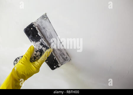 Del lavoratore in mano Guanti gialli holding spatola per applicare una patch a un foro con la spatola con gesso o stucco in parete bianca. Il rinnovamento e il processo di riparazione, ri Foto Stock