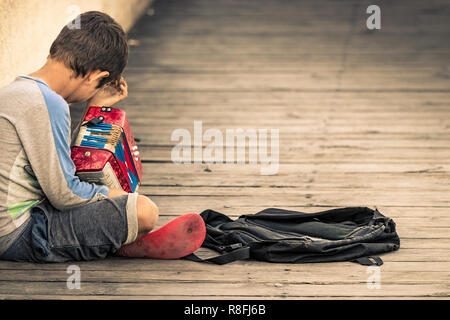 Rhodes, Grecia - Ottobre 10th, 2018: un ragazzo giocatore di fisarmonica seduti all'aperto sul pavimento guardando giù, stanco e triste, di fronte al porto di Rodi, Gr Foto Stock