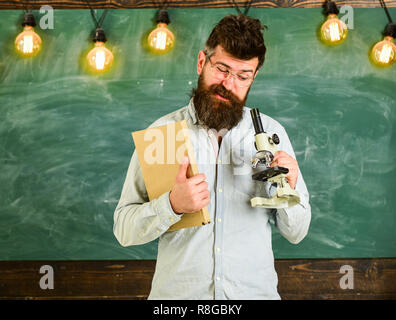 Uomo con barba e baffi sulla faccia di occupato. La ricerca scientifica del concetto. Scienziato detiene il libro e microscopio, lavagna su sfondo. Insegnante in occhiali detiene il libro e microscopio. Foto Stock