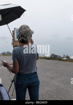 Un artista femminile è in piedi in un parcheggio con un unbrella proteggendo il suo lavoro, pittura foggy acqua scena di fronte a lei. Foto Stock