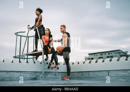 Uomo in possesso di un basket in piedi con due atlete sul tetto scale dopo un allenamento fitness. Basso angolo vista di tre persone fitness un rilassante Foto Stock