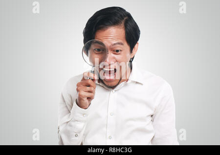 Foto immagine ritratto di un divertente giovane imprenditore asiatico sembrava molto felice, sorpreso e sorridente per trovare qualcosa mentre guardando nella lente di ingrandimento Foto Stock