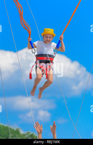 Una ragazzina salta in alto su un trampolino con corde di gomma contro il cielo blu e nuvole bianche. Avventura e Sport Estremi. Concetto di estate vac Foto Stock