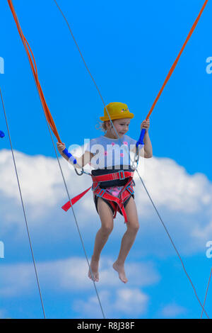 Una ragazzina salta in alto su un trampolino con corde di gomma contro il cielo blu e nuvole bianche. Avventura e Sport Estremi. Concetto di estate vac Foto Stock