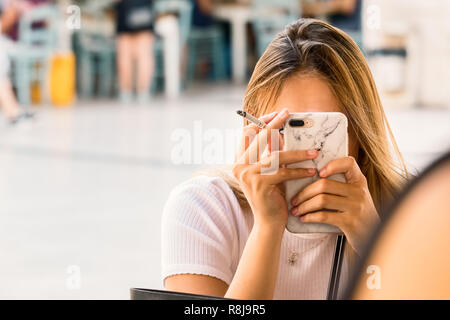 Croazia, Split - Settembre 22th, 2018: una donna bionda si copre la faccia con la sua azienda di telefonia mobile un cigarrete seduti in un caffè all'aperto in Split, C Foto Stock