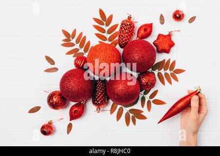 La femmina mano azienda rosso giocattolo ad albero facendo festa laica piana composizione. Donna fare creativo decorazioni di Natale sul tavolo bianco, vista dall'alto. Foto Stock