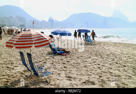 Molti ombrelloni e sedie da spiaggia e molte persone godono di luce del sole mattutina con la vista di Rio de Janeiro del Brasile sullo sfondo Foto Stock