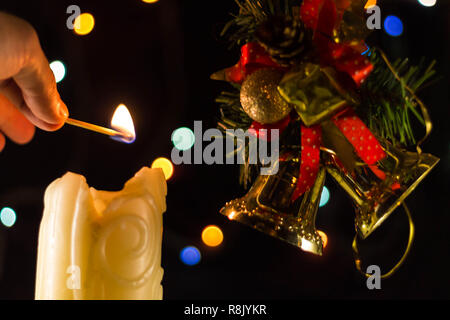 Una scintillante luci di candela una mano con un match. Vicino a dei rami di albero di Natale campane. Foto Stock