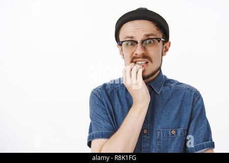 Nervoso e preoccupato in questione di buona cercando maschio con la barba in occhiali trendy beanie e shirt tenendo la mano sul labbro inferiore staring ha scosso e ansiosi in telecamera facendo enormi terribile errore Foto Stock