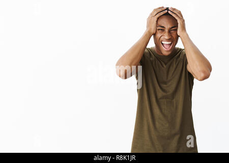 Ritratto di pressioni e di intenso alimentato fino giovane americano africano studente maschio urla dall angoscia tenendo le mani sulla testa in preda al panico e perdere la tempra, infelice permanente ricalcato su sfondo grigio Foto Stock