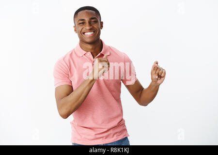 Bello affascinante giovane africano americano guy maschile in rosa polo shirt gesti con le mani come dancing, avendo divertimento sorridente spesa con gioia grande tempo ad amici oltre il muro bianco Foto Stock