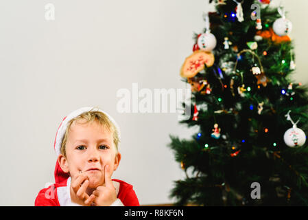 Bello e biondo toddler indossando il costume di Santa accanto ad albero di Natale, isolato sullo sfondo bianco. Foto Stock