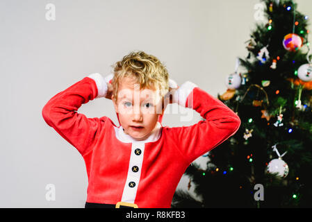 Bello e biondo toddler indossando il costume di Santa accanto ad albero di Natale, isolato sullo sfondo bianco. Foto Stock