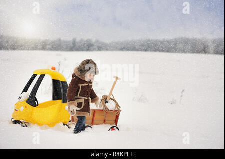 Happy Little Boy in inverno abiti. Il bambino è la riproduzione al di fuori in inverno. Un giallo auto giocattolo e un piccolo carrello riempito con neve, bloccato nella neve Foto Stock