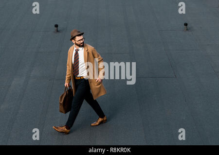 Barbuto imprenditore nel cappotto e hat camminando con la mano in tasca Foto Stock