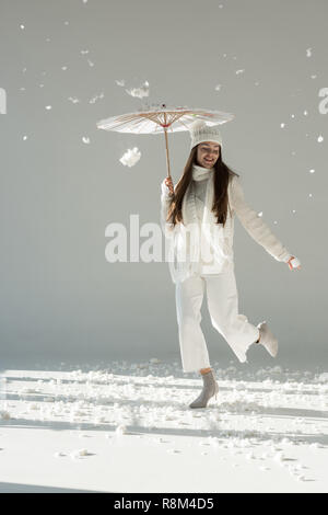 Donna attraente nel quartiere alla moda di inverno maglione e sciarpa jumping in carta ombrello, neve caduta su bianco Foto Stock