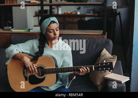 Bellissima gara di misto ragazza in turchese maglione archetto e suonare la chitarra acustica sul divano di casa Foto Stock